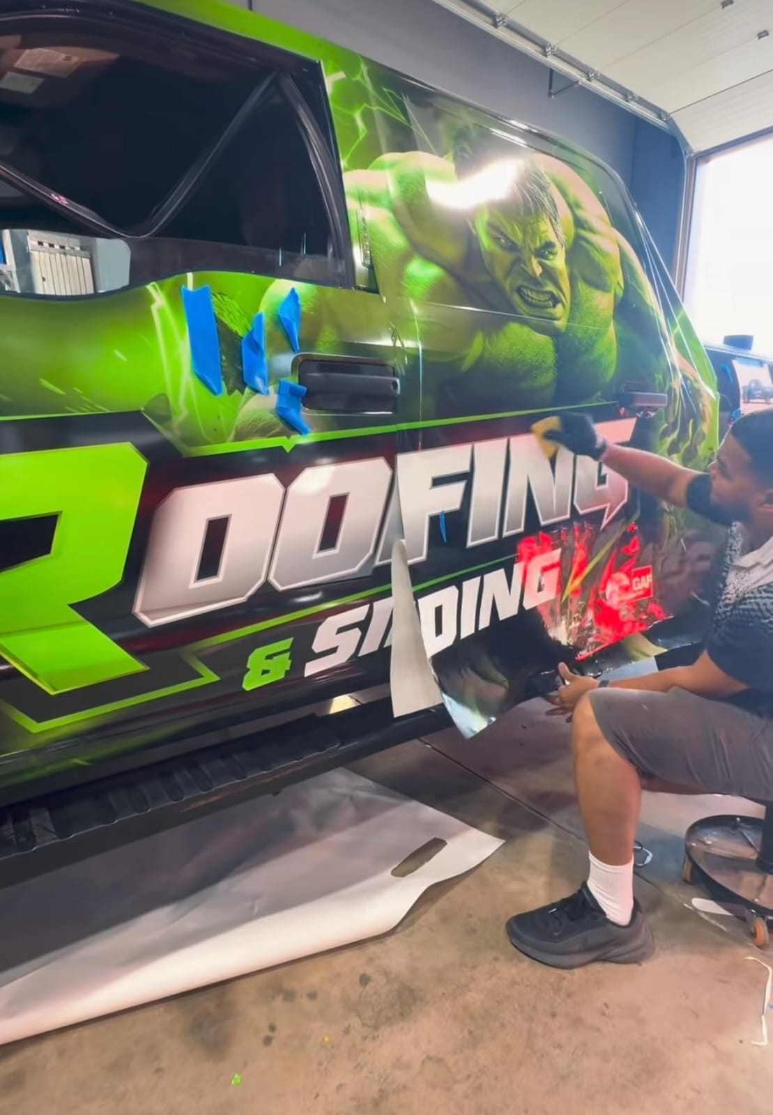 Technician installing a bold green Hulk-themed roofing and siding truck wrap